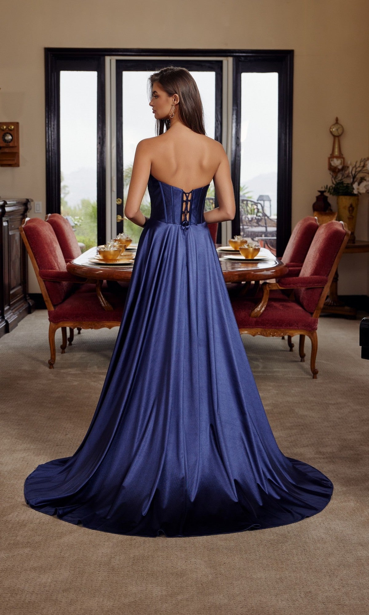 A woman wearing the Nox Anabel K1735 long prom dress, featuring a strapless satin bodice with lace-up corset and flowing A-line skirt, stands facing away in an elegant dining room.